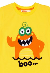 Gul bomulds t-shirt med et orange monster med grønne øjne og et stort smil, der vinker med teksten "boo..." i sort nedenfor.