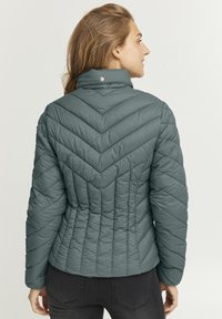 Padded jacket v tlmenom tealovom odtieni, so vzorom v tvare píly, vysokým golierom a patentným zapínaním na krku. Hladká textúra, priliehavý strih.