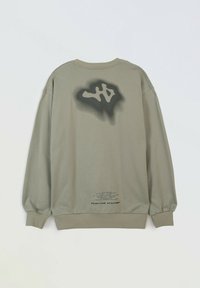 Jasno oliwkowy sweatshirt z abstrakcyjnym czarnym wzorem w sprayu w okolicy górnej części pleców oraz małym czarnym tekstem blisko dolnego hemu.