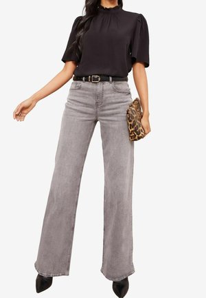 Kobieta ubrana w czarną bluzkę z bufkami, wpuszczoną w szare, szerokie dżinsy z wysokim stanem, z czarnym paskiem, trzymająca kopertówkę w panterkę.