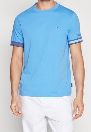 Homme portant un t-shirt bleu ciel avec poignets de manches rayés, petit logo sur la poitrine et pantalon blanc, debout devant un fond uni.