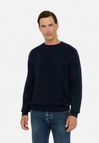 Granatowy sweter dziewiarski z teksturowanym wzorem, z prążkowanymi mankietami i dolnym brzegiem, z kolorowymi flekami wplecionymi w materiał. Noszony z niebieskimi dżinsami.