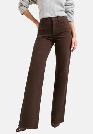 Donna che indossa pantaloni marroni a vita alta e ampia gamba abbinati a un maglione grigio chiaro, in posa con una mano sui fianchi.