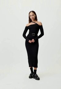 Črna rebrasta pletena obleka z off-shoulder dizajnom, dolgimi rokavi in detajlom pasu, v kombinaciji s črnimi čevlji in nogavicami.