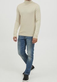 Beige Rollkragenpullover aus weichem Material, kombiniert mit blauen Jeans und schwarzen Sneakers, vor einem weißen Hintergrund stehend.