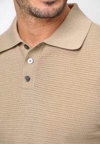 Beige stickad piké med kort krage, prydd med ett texturerat mönster, två knappar i mörkgrått och en avslappnad passform.