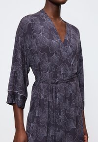 Vestito avvolgente grigio scuro con leggere fantasie floreali, maniche a tre quarti e una cintura di tessuto legata in vita, indossato da una persona con pelle scura.