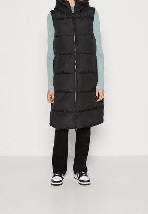 Frau trägt eine lange schwarze gesteppte ärmellose Kapuzenweste, hellblauen gerippten Pullover, schwarze Hose und schwarz-weiße Sneakers.