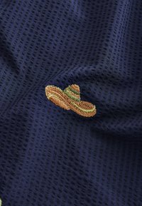Petit sombrero brodé marron et vert sur tissu bleu foncé texturé.