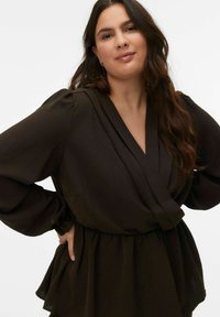 Bruine, gestructureerde blouse met een diepe V-hals, lange gebolde mouwen en een elastische taille voor een losse pasvorm. Geen zichtbare patronen of accessoires.