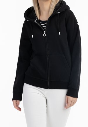 Frau trägt eine schwarze Kapuzenjacke mit Reißverschluss und Kordeln über einem gestreiften Hemd und weißer Hose, stehend vor einem weißen Hintergrund.
