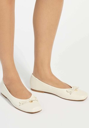 Jambes portant des ballerines blanches avec un petit nœud et un charme doré sur un fond blanc.