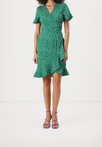 Robe portefeuille verte à motif floral blanc, manches courtes à volants et ourlet évasé. Portée avec des sandales à lanières violettes.