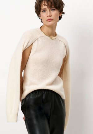 Beige gebreide trui met een open schouderontwerp, lange mouwen, zachte textuur, gecombineerd met hoog opgesneden zwarte leren broek.