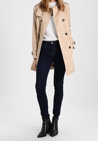 Beige trenssi takki, jossa on kaksiriviset napit, hartiahihnat, valkoisen topin päällä, tummien kapeiden farkkujen ja mustien nilkkureiden kanssa.