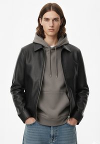 Giacca di pelle nera con collo, indossata sopra una felpa grigia con cappuccio. Modello con capelli lunghi e jeans blu. Dettaglio cerniera sulla giacca.