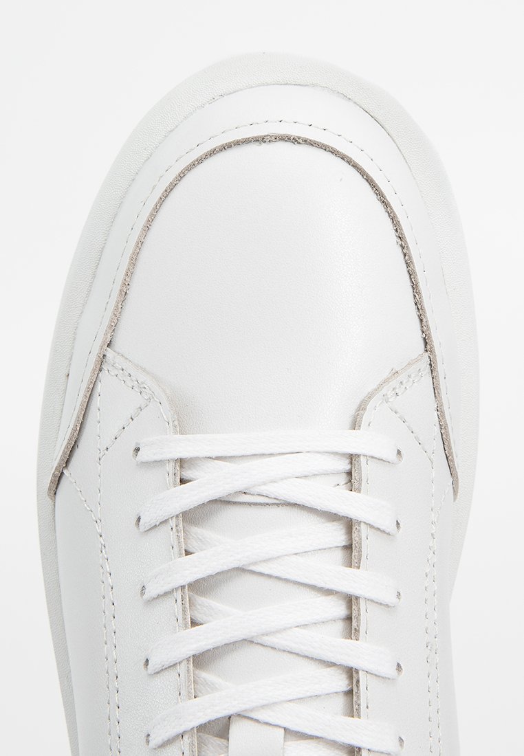 Baskets blanches avec une tige en cuir lisse, lacets plats, couture renforcée et bout arrondi. Accents texturés près de la semelle.