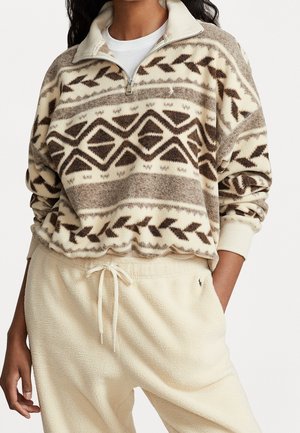 Mujer con un forro polar beige y marrón con estampado, media cremallera, y pantalones de forro polar color crema con cintura ajustable con cordón.