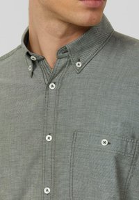 Overhemdbloes met kraag, lichtgrijze stof met subtiele textuur, witte knopen, een borstzak en gestikte details langs de naden.