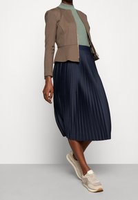 Personne portant une veste ajustée marron, un haut côtelé vert, une jupe midi plissée bleu marine et des baskets beiges, se tenant contre un fond blanc.