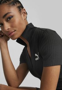 Svart ribbestrikket polo-skjorte med en tettsittende design, med høy krage og hvit Puma-logo på venstre bryst. Glatt stofftekstur.