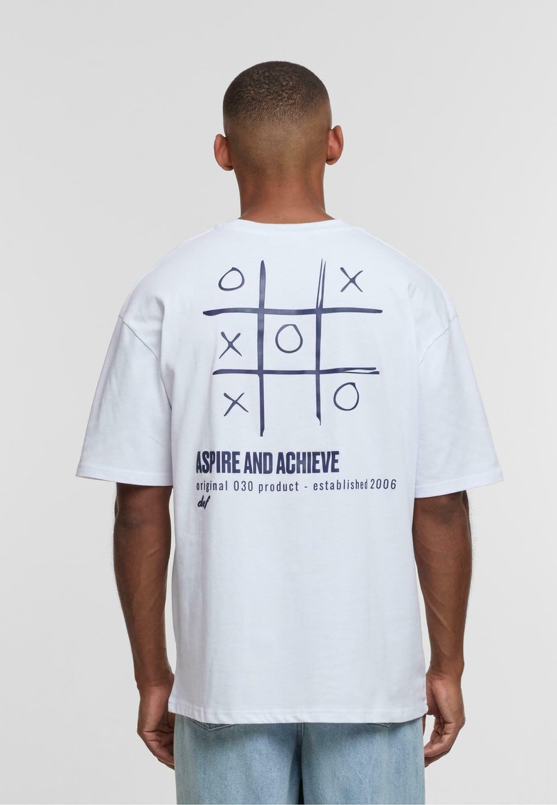 White cotton t-shirt featuring a tic-tac-toe graphic in dark blue on the back, with text reading "ASPIRE AND ACHIEVE" below. Loose fit.