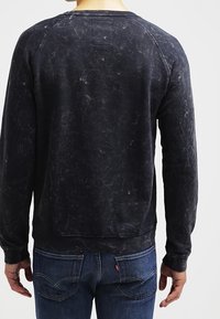 Felpa nera effetto consumato con scollo rotondo, maniche raglan e polsini a costine. Indossata con jeans blu caratterizzati da un logo discreto sulla tasca.