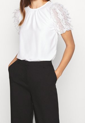 Vrouw draagt een witte blouse met doorschijnende gerimpelde mouwen en zwarte broek met hoge taille, handen in de zakken tegen een lichte achtergrond.