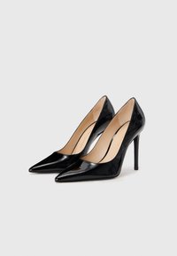 Tacones altos de charol negro con punta puntiaguda y diseño elegante. Tacón de aguja y forro interior beige con un detalle sutil de la marca.