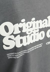 Hallikas puuvillane t-särk, millel on suured valged sõnad "Original Studio", mille all on kirjatud väiksemaid sõnu.