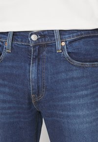 Tmavě modré džínové kalhoty klasického střihu, s měděným kováním, předními kapsami a kontrastním prošíváním podél švů.