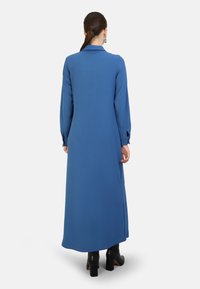 Long-sleeve, blue dress made of smooth fabric, featuring a collar, side slits, and button details at the cuffs. Worn with black heeled boots.