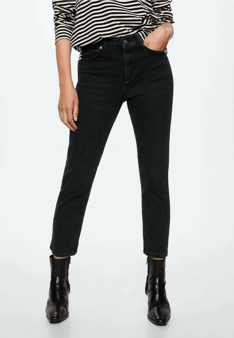 Personne portant un jean slim noir taille haute, un haut rayé à manches longues et des bottines en cuir noir, debout avec une main dans la poche.