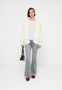 Γυναίκα που φοράει λευκό πουλόβερ με πλέξη καλώδιο και ζακέτα, ανοιχτό μπλε τζιν με καμπάνα, μπορντό παπούτσια, κρατώντας μικρή μαύρη τσάντα, στέκεται μπροστά σε λευκό φόντο.