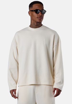 Mann trägt cremefarbenes langärmeliges Oberteil und passende Hose, schwarze Sonnenbrille, mit Hals- und Handtattoos, steht vor einem einfarbigen Hintergrund.