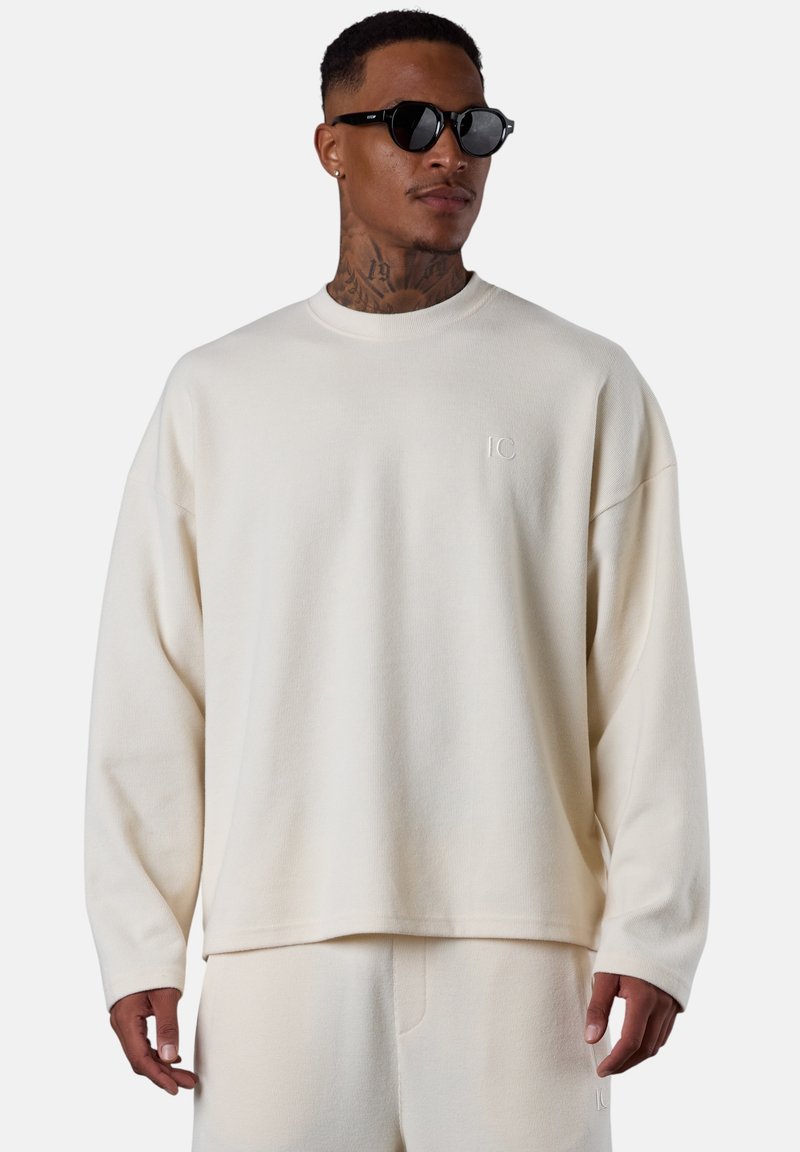 Man wearing cream-colored long-sleeve top and matching pants, black sunglasses, with neck and hand tattoos, standing against a plain background.