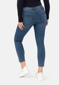 Schmale Jeans aus dunkelblauem Denim, mit mittelhoher Taille, zwei Gesäßtaschen und einem ausgefransten Saum. Kombiniert mit weißen Sneakers.