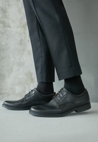 Chaussures de ville en cuir noir avec une finition lisse, des détails de laçage, un talon bas, et associées à un pantalon noir et des chaussettes sur une surface texturée.