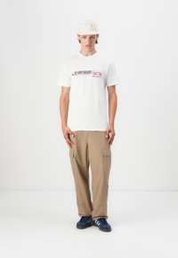 Hvit t-skjorte med trykt tekst og grafikk, beige cargo-bukser, og marineblå sneakers med lyseblå striper, brukt med en lys beige caps.