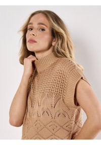 Pletený top bez rukávů ve světle béžové barvě s vysokým límcem. Obsahuje otvorový vzor a geometrický design na lemu. Měkká textura.