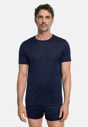 Mies, jolla on lyhyet kiharat hiukset ja parta, pukeutuneena tummansiniseen lyhythihaiseen puseroon ja sitä vastaaviin shortseihin, seisoo vaaleaa taustaa vasten.