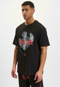 Čierne tričko s krátkym rukávom, ktoré má grafický potlač postavy s anjelskými krídlami a textom "Thug Life" v červenej farbe, umiestnené na svetlom pozadí.