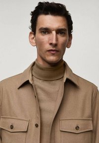 Tan wollen shirtjacket met twee knoopzakken op de borst, gelaagd over een bijpassende tan coltrui. Gladde textuur en getailleerde pasvorm.