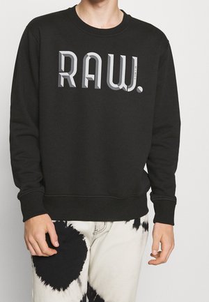 Persona che indossa una felpa nera con la scritta "RAW." in grassetto grigio e pantaloni bianchi con grandi motivi circolari neri.