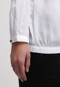 Vit långärmad blus med en insydd elastisk nederkant och knappade manschetter. Tyget verkar lätt och slätt, med en enkel design.