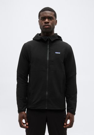 Νέος άνδρας που φοράει μαύρο μπουφάν με κουκούλα και φερμουάρ, με ένα μικρό λογότυπο Patagonia στο στήθος, στέκεται με τα χέρια χαλαρά δίπλα στο σώμα του.