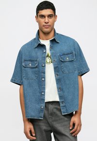 Ljusblå kortärmad denimskjorta med två framfickor, knapp-up design och klassisk krage, bärs över en vit grafisk t-shirt.