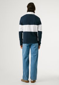 Mørkeblå langærmet polo med en hvid vandret stribe, parret med lyseblå lige ben jeans og brune sko, vist fra ryggen.