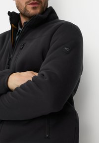 Zwarte fleecejas met een hoge kraag, een getextureerde voering aan de binnenkant, zakken met rits en een logopatch op de linkermouw.