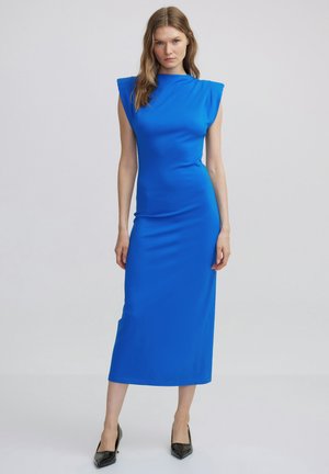 Blue, form-fitting midi dress with shoulder pads, high neckline, and smooth texture. Accented with black, pointed-toe heels.